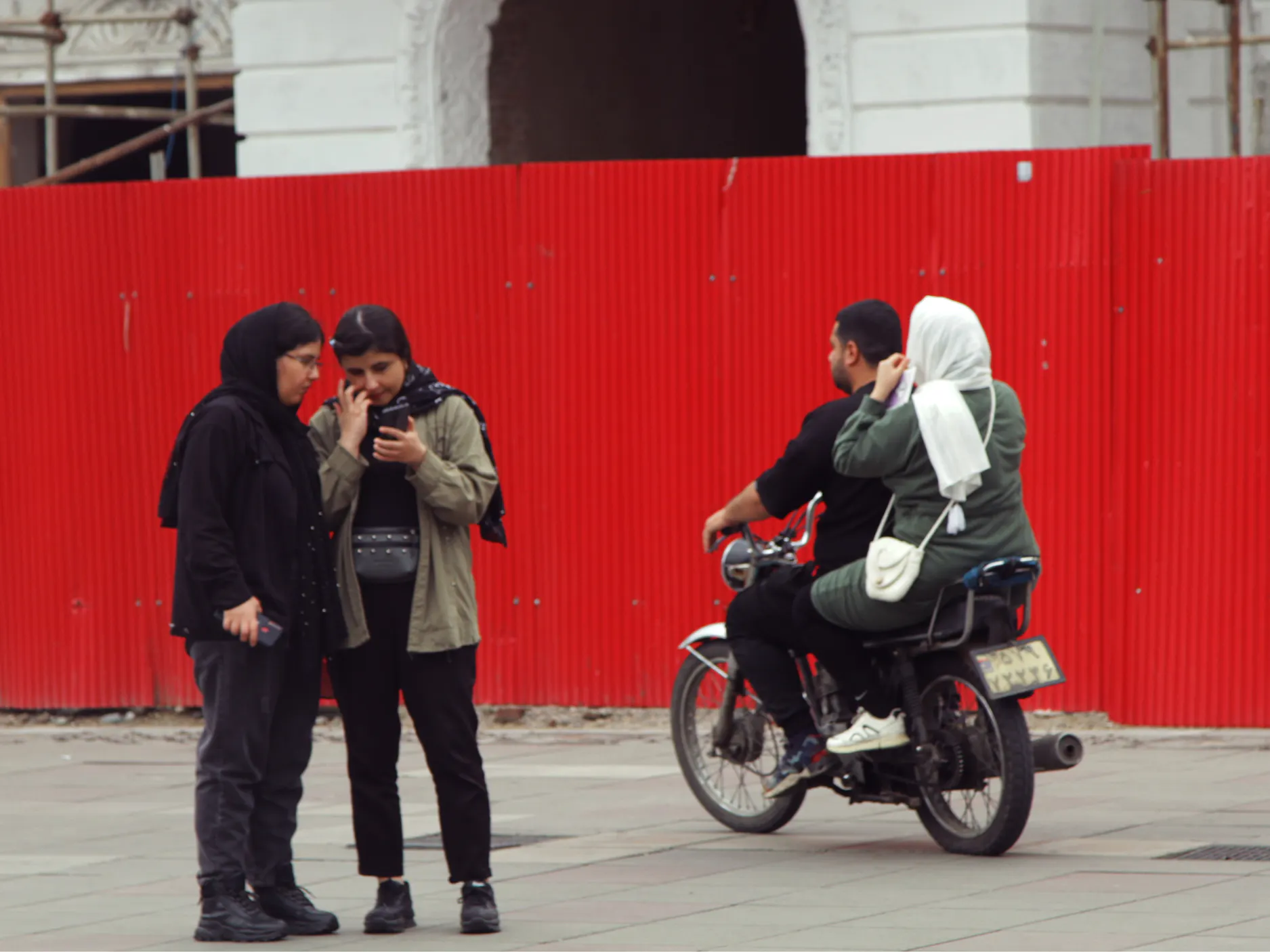 Iranska ulica, dve ženski gledata v telefon, mimo se vozita moški in ženska na motorju